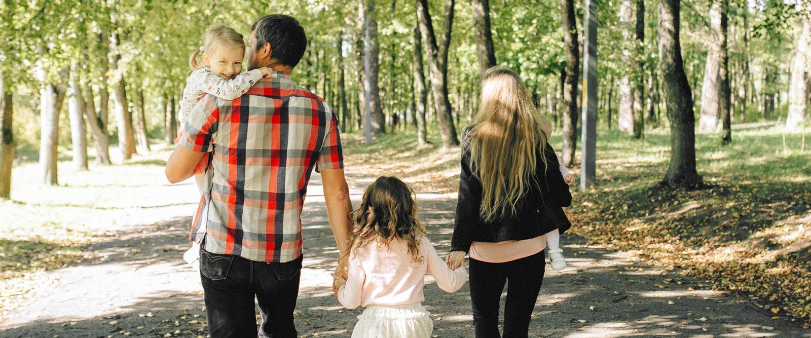 木もれ日の公園の道を、子どもと手をつないで歩いていく家族の後ろ姿